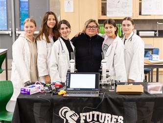 5 students in lab coats are posed with a staff member in front of their display